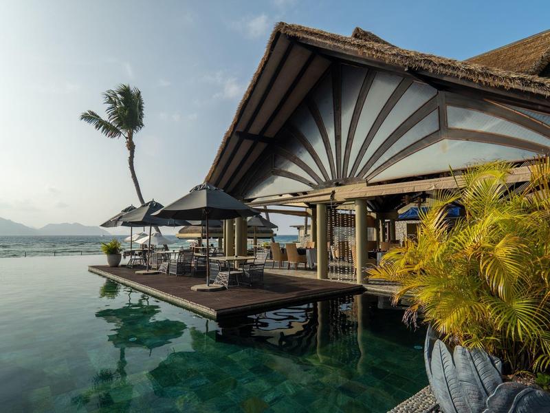 Luxury overwater villa with infinity pool, palm trees, and panoramic mountain view at sunrise.