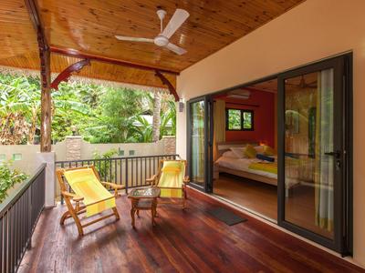 Covered wooden terrace with two lounge chairs, table, and view into a bedroom through sliding glass door.