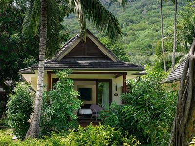 Kleines Haus umgeben von üppigem Grün und Palmen vor einem bewaldeten Hügel.