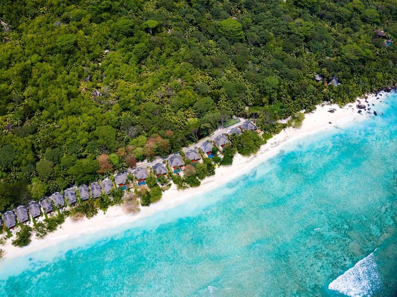 Vista aérea de un resort en la playa con cabañas en la arena blanca junto a agua turquesa y densa selva.
