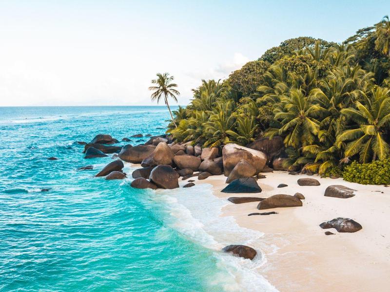 Weißer Sandstrand mit großen Felsen und Palmen am türkisblauen Ozean.