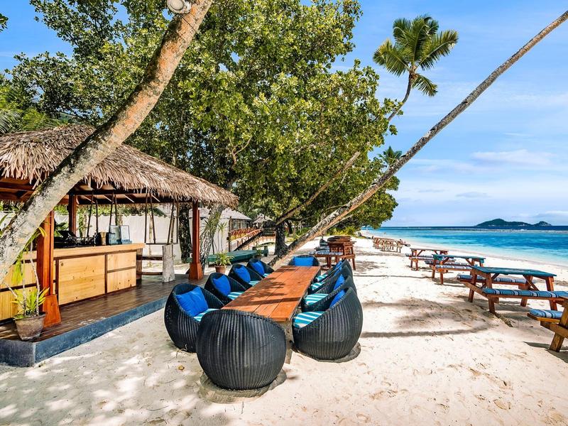 Strandrestaurant mit Tischen, Stühlen und Blick auf das türkisfarbene Meer und den blauen Himmel.