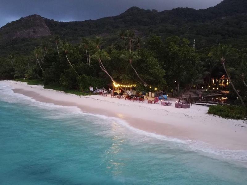 Strand mit weißem Sand, türkisfarbenem Wasser und beleuchtetem Resort am Fuß eines bewaldeten Hügels bei Dämmerung.