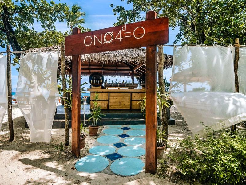 Strandbar mit hölzernem Eingangsschild und türkisfarbenen Steinen auf Sandboden.