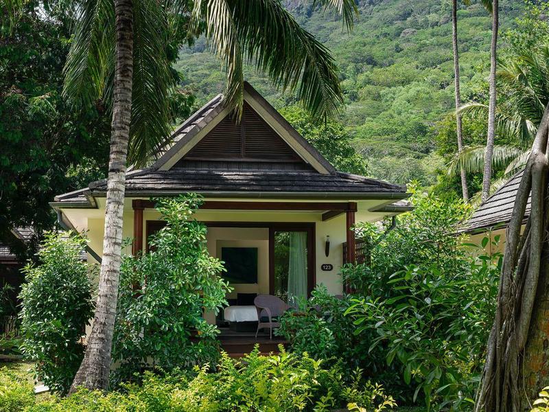 Kleines Haus umgeben von üppigem Grün und Palmen vor einem bewaldeten Hügel.