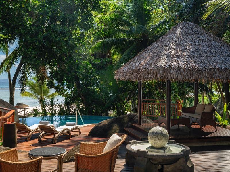 Outdoor-Lounges mit Stühlen, Tischen und einem Strohdach-Pavillon neben einem Pool und tropischem Garten.