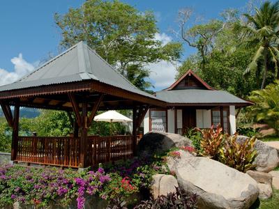 Holzhütte mit Veranda und Garten mit großen Steinen, Blumen und Palmen unter blauem Himmel.