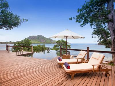 Lounge-Bereich mit Holzboden, Liegen, Sonnenschirm und Blick auf Berge und See im Hintergrund.