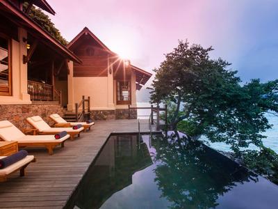 Luxuriöse Holzterrasse mit Liegestühlen, Pool und Baum vor einem Haus am Wasser bei Sonnenuntergang.