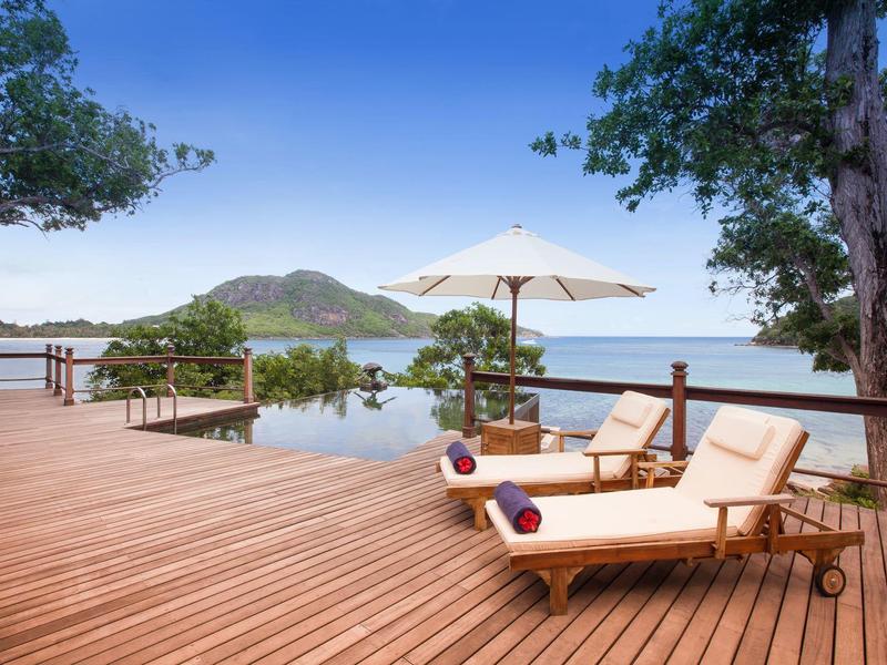 Lounge-Bereich mit Holzboden, Liegen, Sonnenschirm und Blick auf Berge und See im Hintergrund.