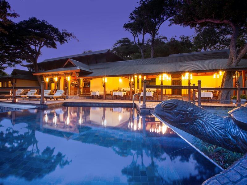 Ein großes, beleuchtetes Haus spiegelt sich im blauen Wasser eines Pools mit Schildkrötenfigur wider.