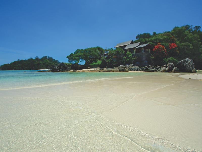 Weißer Sandstrand mit klarem Wasser, grünen Bäumen und einem Gebäude am Rand unter blauem Himmel.