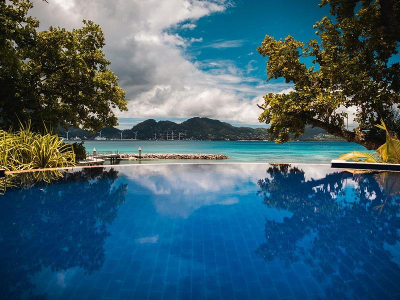 A pool overlooking a beach and forested mountains under a cloudy sky.