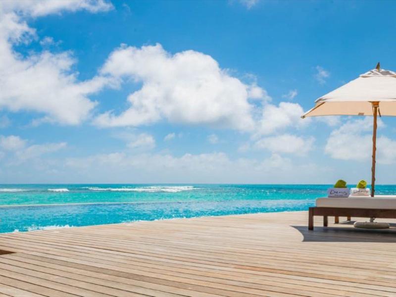 Holzdeck mit Liegestuhl und Sonnenschirm am türkisfarbenen Meer unter blauem Himmel mit Wolken.