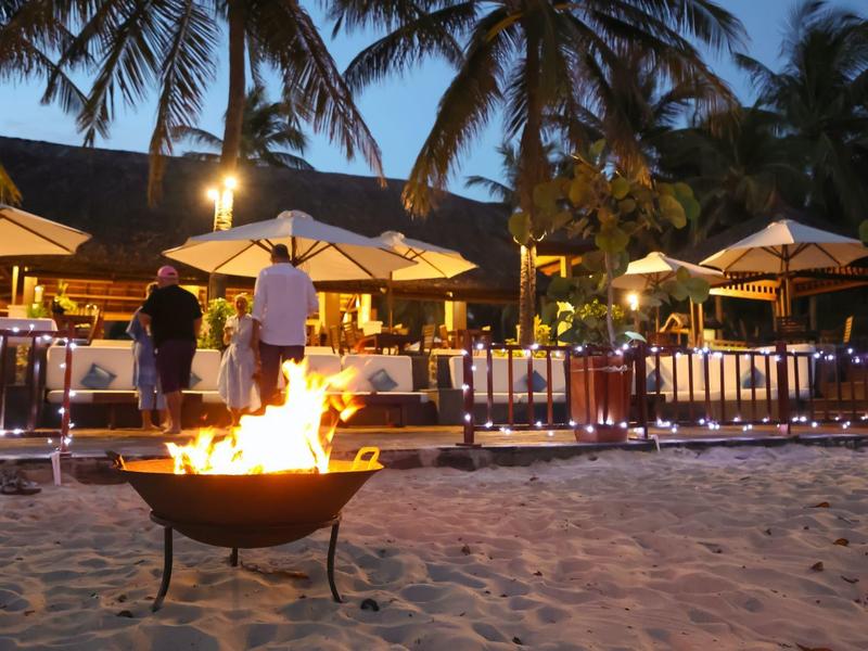 Strand mit Feuerschale, Palmen und beleuchtetem Restaurant im Abendlicht.