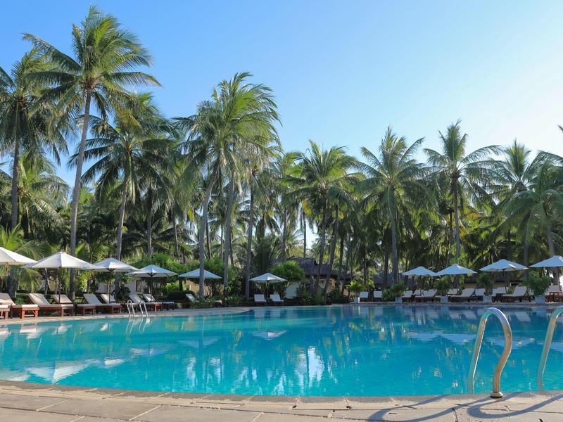 Großer Pool mit klarem Wasser, umgeben von Palmen und Liegestühlen unter Sonnenschirmen.