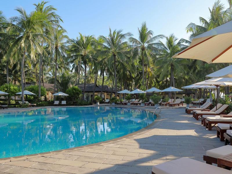 Großer Swimmingpool mit Holzdeck, Liegestühlen und Sonnenschirmen, umgeben von Palmen.