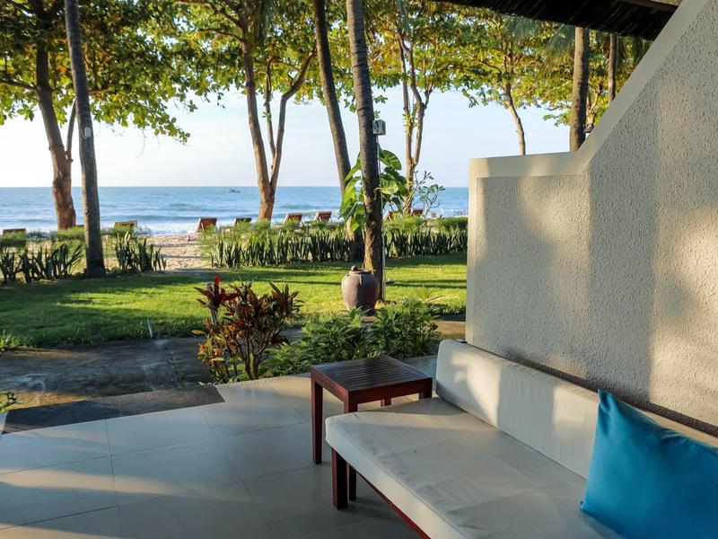 Terrasse mit weißem Sofa und blauer Kissen, Blick auf grüne Bäume und Meer im Hintergrund.
