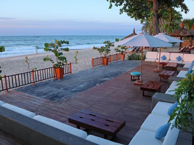 Terrasse mit Holzmöbeln und Sonnenschirmen neben einem Sandstrand und ruhigem Meer unter blauem Himmel.