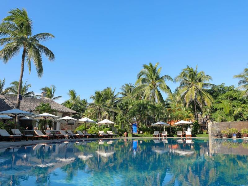 Großer Pool mit klar blauem Wasser, umgeben von Palmen und Liegestühlen unter Sonnenschirmen.