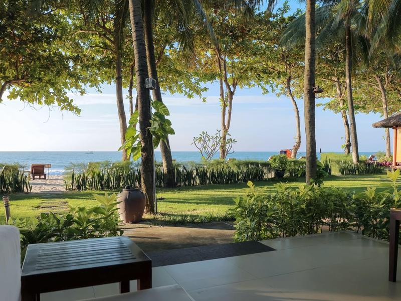 Grüner Garten mit Palmen, Bänken und Blick auf Meer unter blauem Himmel am Strand.