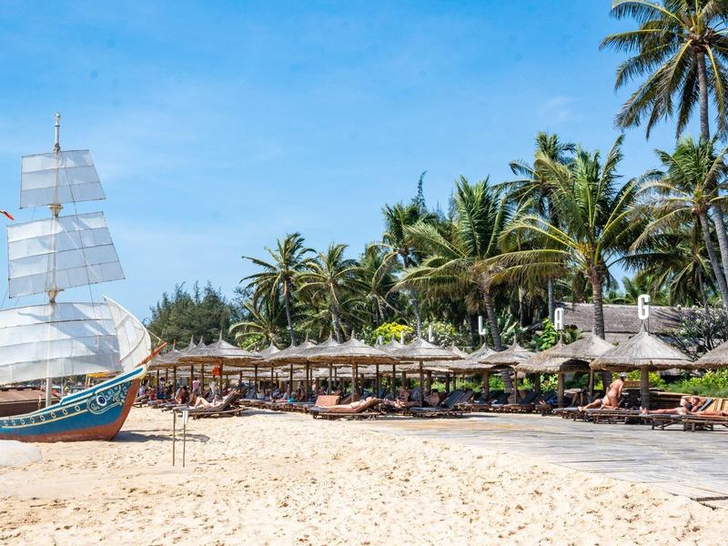 Pláž s pískem, palmami, lehátky a velká plachetnice pod modrou oblohou.