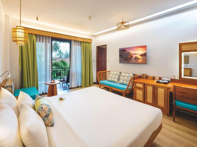 Světý hotelový pokoj s velkou postelí, pohovkou, psacím stolem a balkonem s výhledem na zeleň.