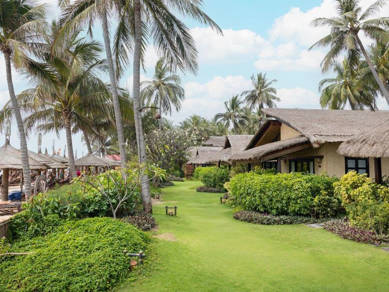 Zelená zahrada s bungalovy a palmami blízko pláže pod modrou oblohou.