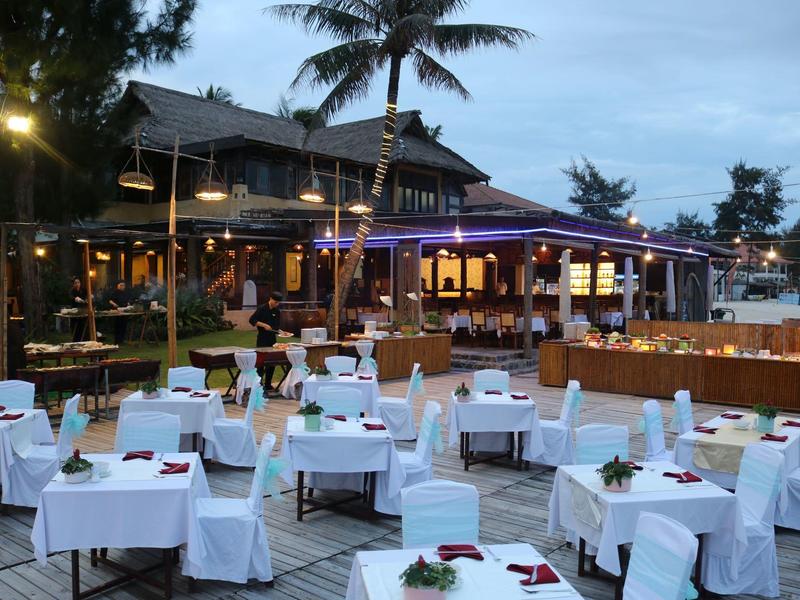 Venkovní restaurace s bílými ubrusy na stolech a dřevěnou chatkou v pozadí za soumraku.