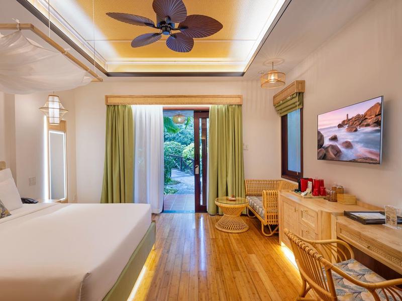 Moderní hotelový pokoj s velkou postelí, stropním ventilátorem, psacím stolem a výhledem na terasu s zelení.