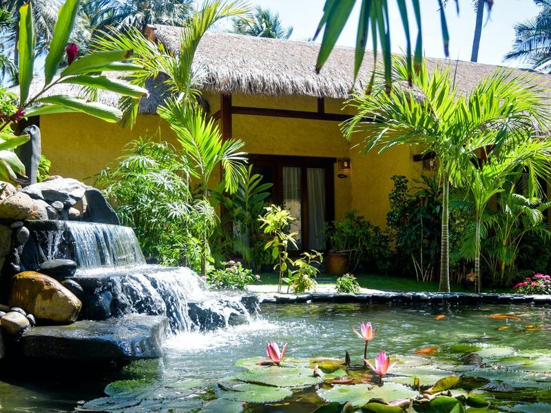 Tropická zahrada s vodopádem a jezírkem před žlutým domem se doškovou střechou.