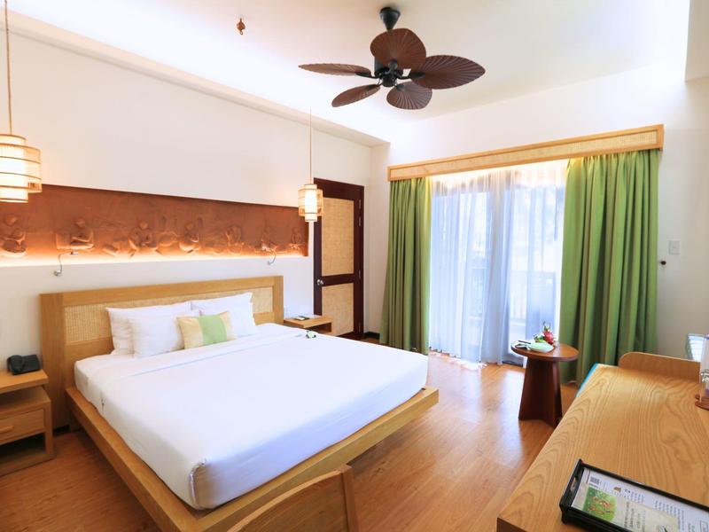 Světý hotelový pokoj s velkou postelí, zelenými závěsy a stropním ventilátorem.