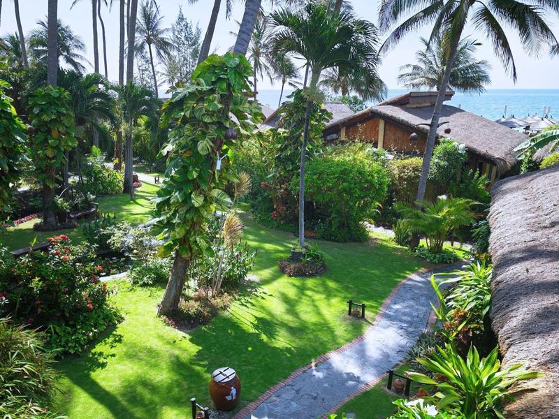 Bujná tropická zahrada s palmami, dlážděnou cestou a dřevěnými domy v pozadí