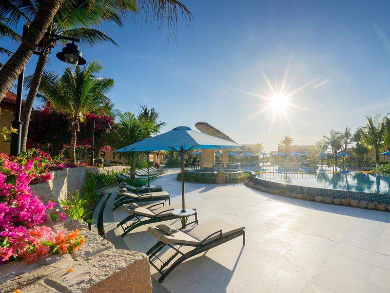 Zonnig hotelzwembad met ligstoelen, parasols en palmbomen.