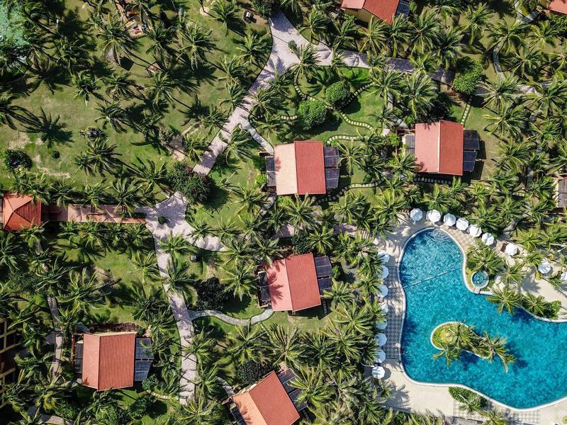 Luchtfoto van bungalowhutten met rode daken en een zwembad omgeven door palmbomen.