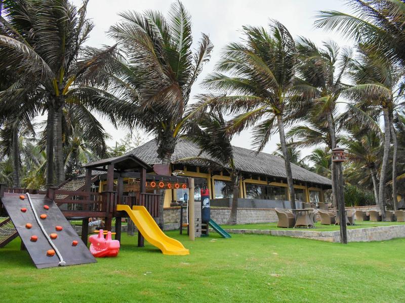 Speelplaats voor kinderen met glijbaan en klimwand voor een gebouw met palmendak.
