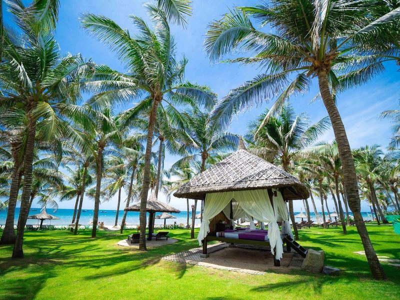 Paviljoen met witte gordijnen tussen palmbomen op een strand onder een blauwe lucht.