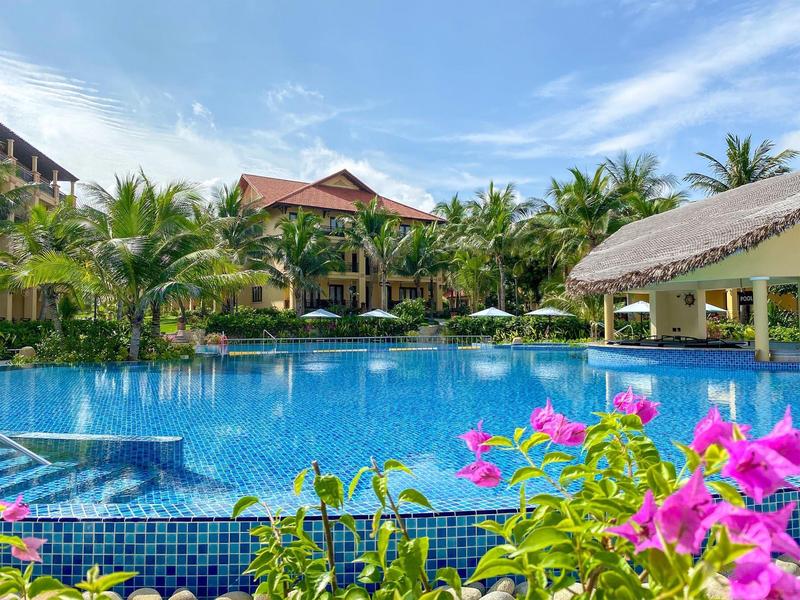 Groot hotelzwembad met bloeiende planten, parasols en palmbomen onder een blauwe lucht.