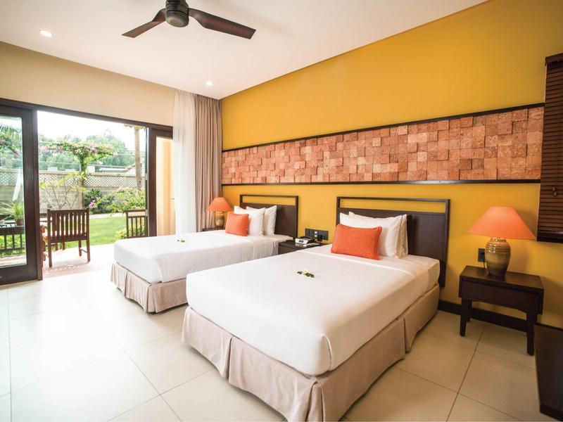 Hotelkamer met twee eenpersoonsbedden, gele muur en balkon met uitzicht op groen.