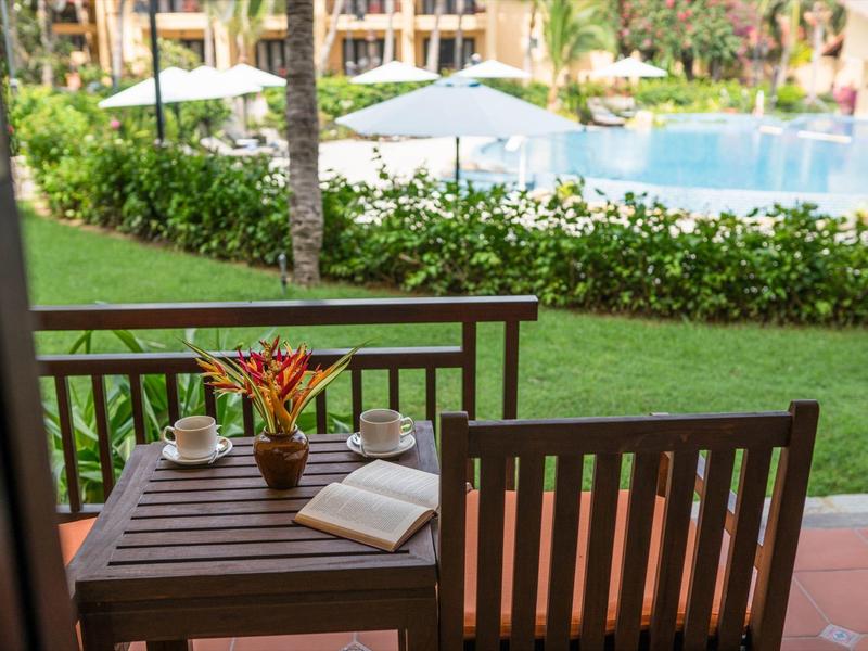 Een houten tafel met een kopje, boek en bloemenvaas op een balkon met uitzicht op het zwembad.