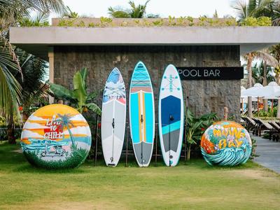 Drei bunte Surfbretter und zwei große bunte Bälle vor einer Poolbar mit Palmen und Liegestühlen.