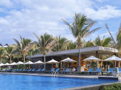 Duży basen z białymi parasolami i palmami przed nowoczesnym budynkiem pod zachmurzonym niebem.