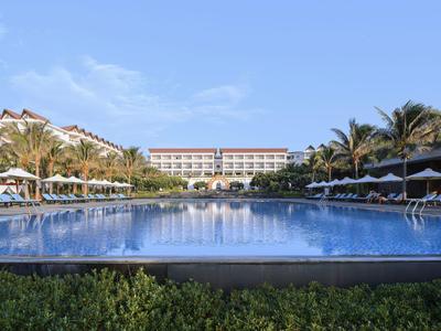 Duży basen przed ośrodkiem z leżakami i palmami pod czystym niebem.