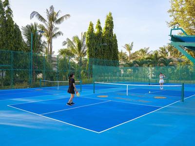 Dwie osoby grają w tenisa na niebieskim boisku na świeżym powietrzu otoczonym drzewami i zielonymi ogrodzeniami.