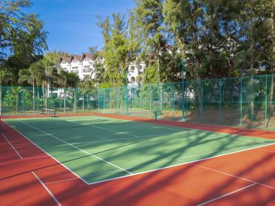 Kort tenisowy z zielonym polem gry i czerwoną krawędzią, otoczony drzewami i ogrodzeniem.