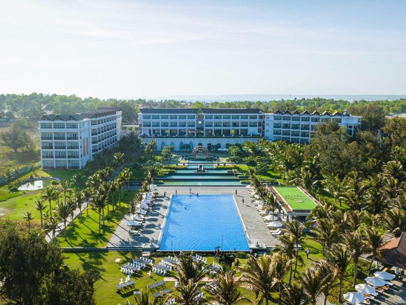Duży hotel z centralnym basenem, otoczony palmami i zielonym krajobrazem.