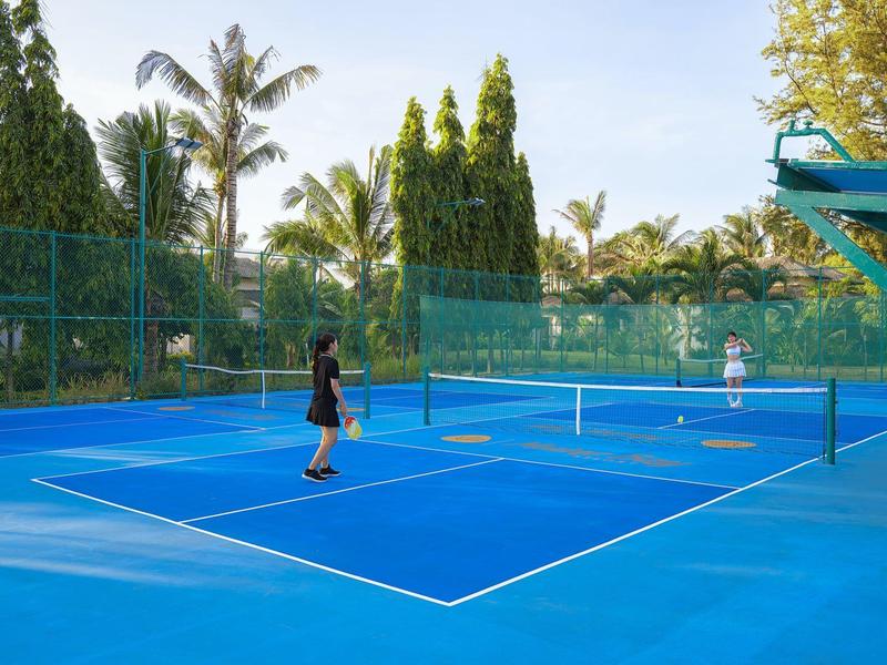 Dwie osoby grają w tenisa na niebieskim odkrytym korcie otoczonym drzewami pod słonecznym niebem.