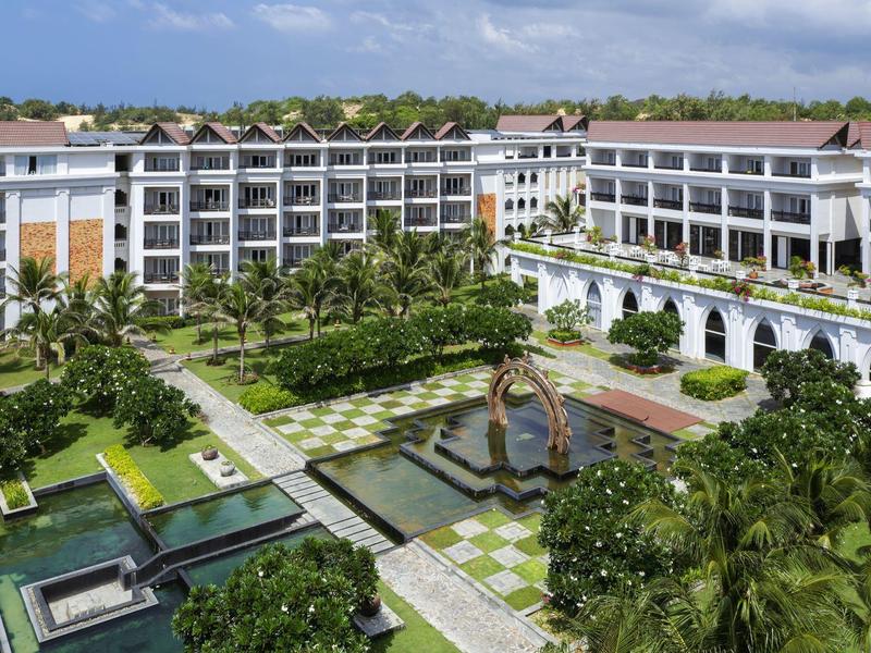 Duży hotel z dziedzińcem, fontanną i bujnym ogrodem pod błękitnym niebem.