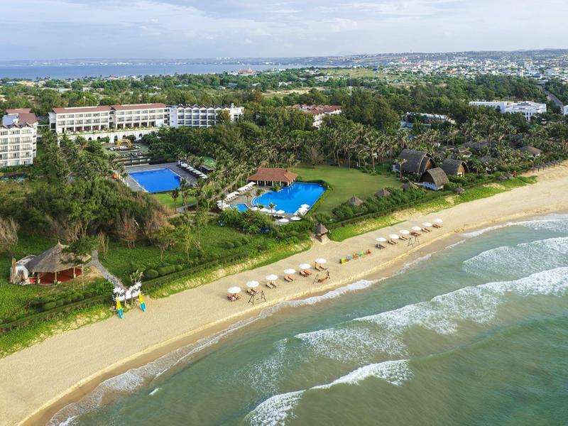 Hotel przy plaży z dwoma niebieskimi basenami, leżakami na piaszczystej plaży i bujną zielenią.