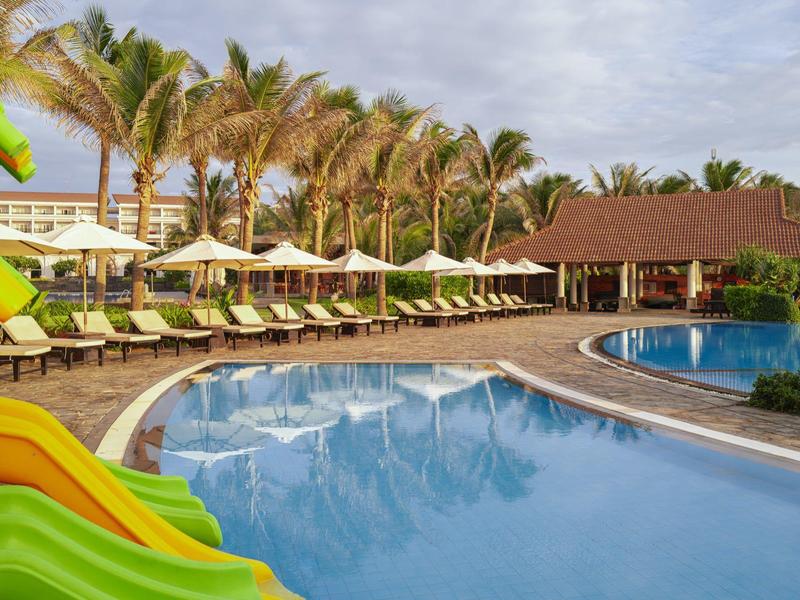 Basen hotelowy z zjeżdżalnią wodną, leżakami i palmami pod pochmurnym niebem.